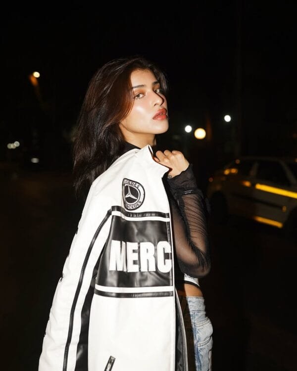 A close-up of a woman holding a white Mercedes-Benz racing jacket over her shoulder, wearing a black sheer top, against a nighttime urban backdrop.