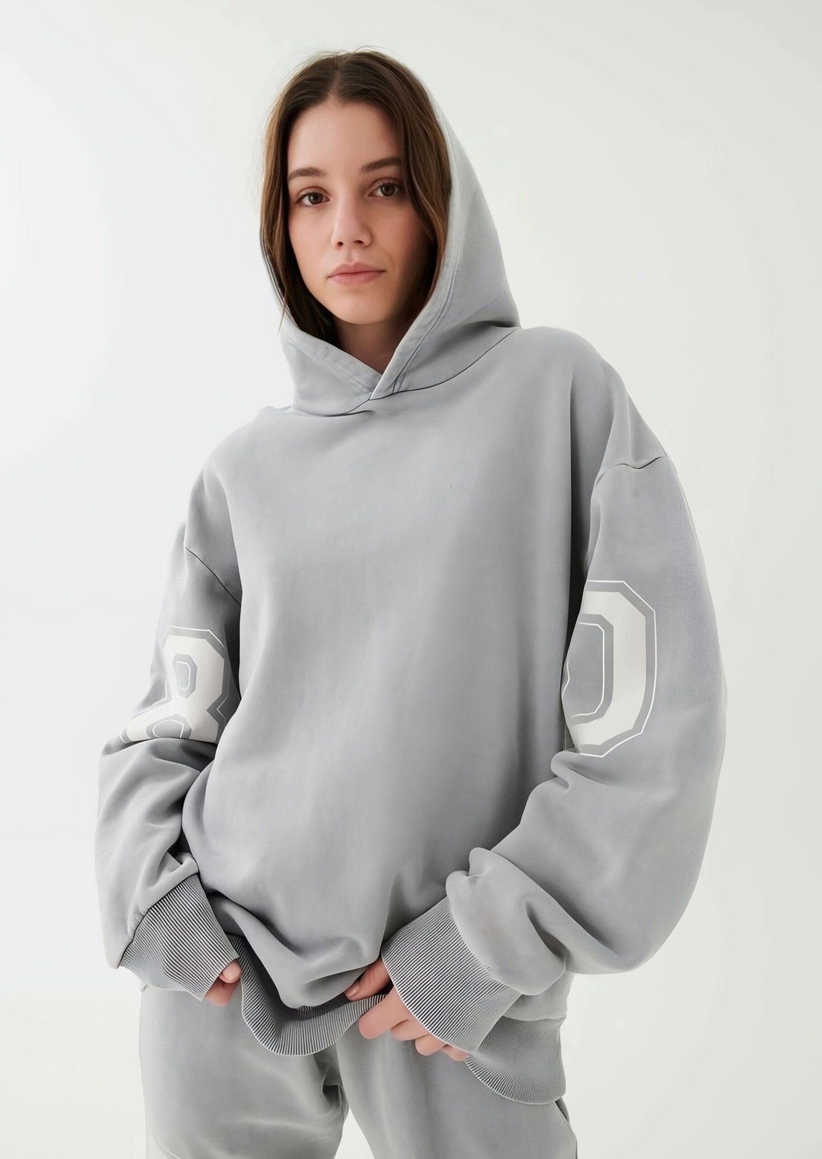 A female model dressed in an zerodas oversized grey hoodie with a subtle textured design on the sleeves. She pairs it with matching grey sweatpants, posing in a relaxed manner against a soft, neutral background.