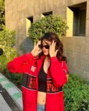 Woman wearing a red Ferrari racing jacket with sunglasses, posing in sunlight near a modern stone wall and greenery.