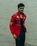 Man standing at the edge of a pier in a red Ferrari jacket, smiling slightly, with various F1 sponsor logos visible.