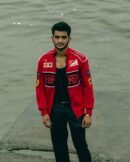 A man in a red Ferrari racing jacket posing confidently by the water, with full front view displaying Ferrari, Shell, and Puma logos.